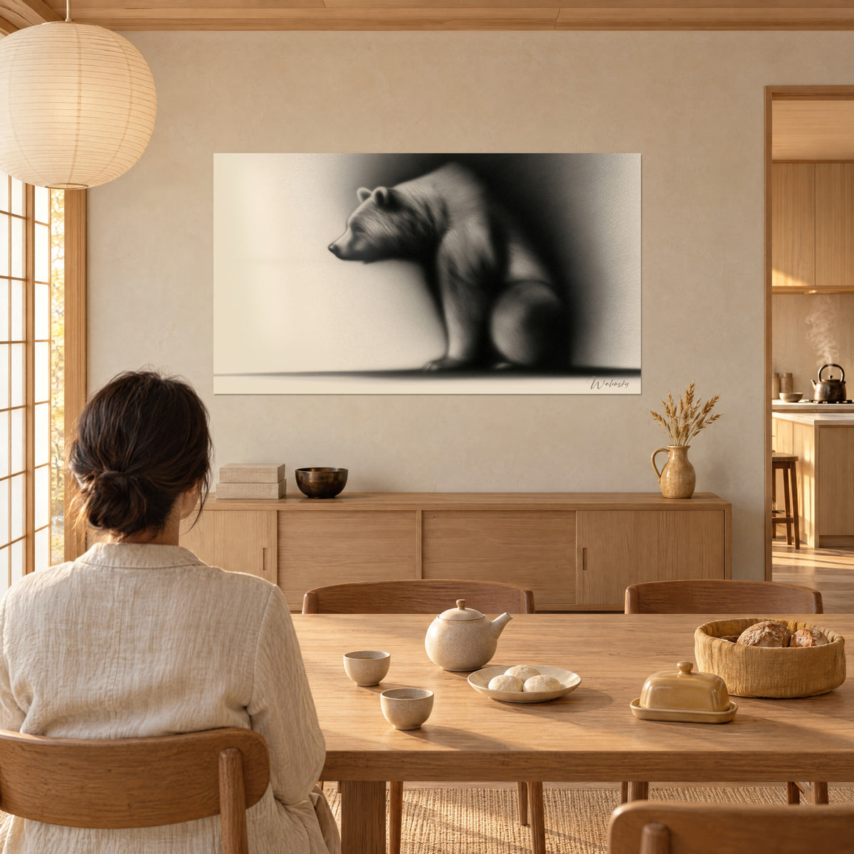 Wall art of a seated bear cub with soft blurred outlines, deep black and cream beige, gazed at by a woman seen from behind seated at a wooden dining nook
