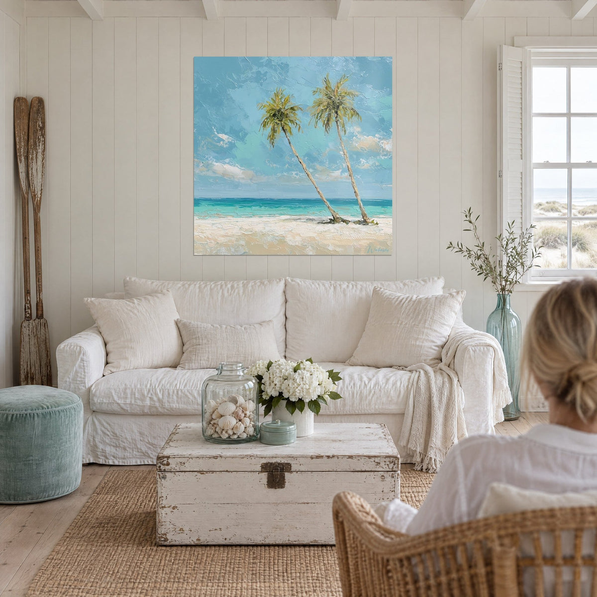 Wall art two palm trees leaning over a turquoise sea, thick-textured blue sky, cream sand, two people seen from behind above a sofa in a bright living room