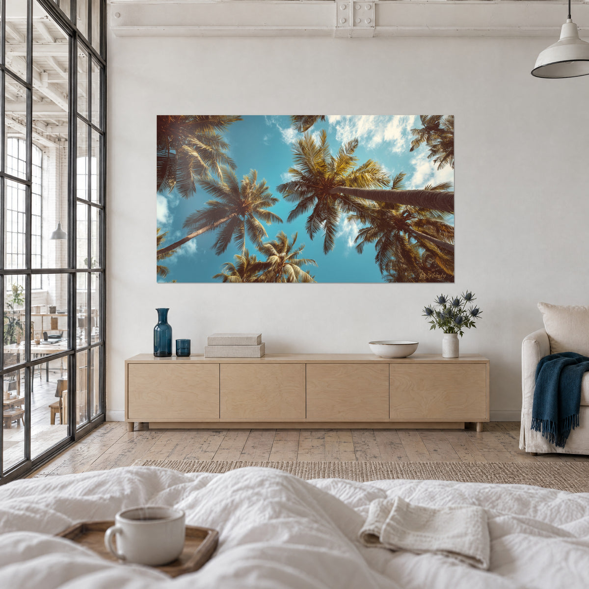 Wall art palm canopy under a turquoise sky, white clouds and brown leaves, couple seen from behind above a bed in a light bedroom