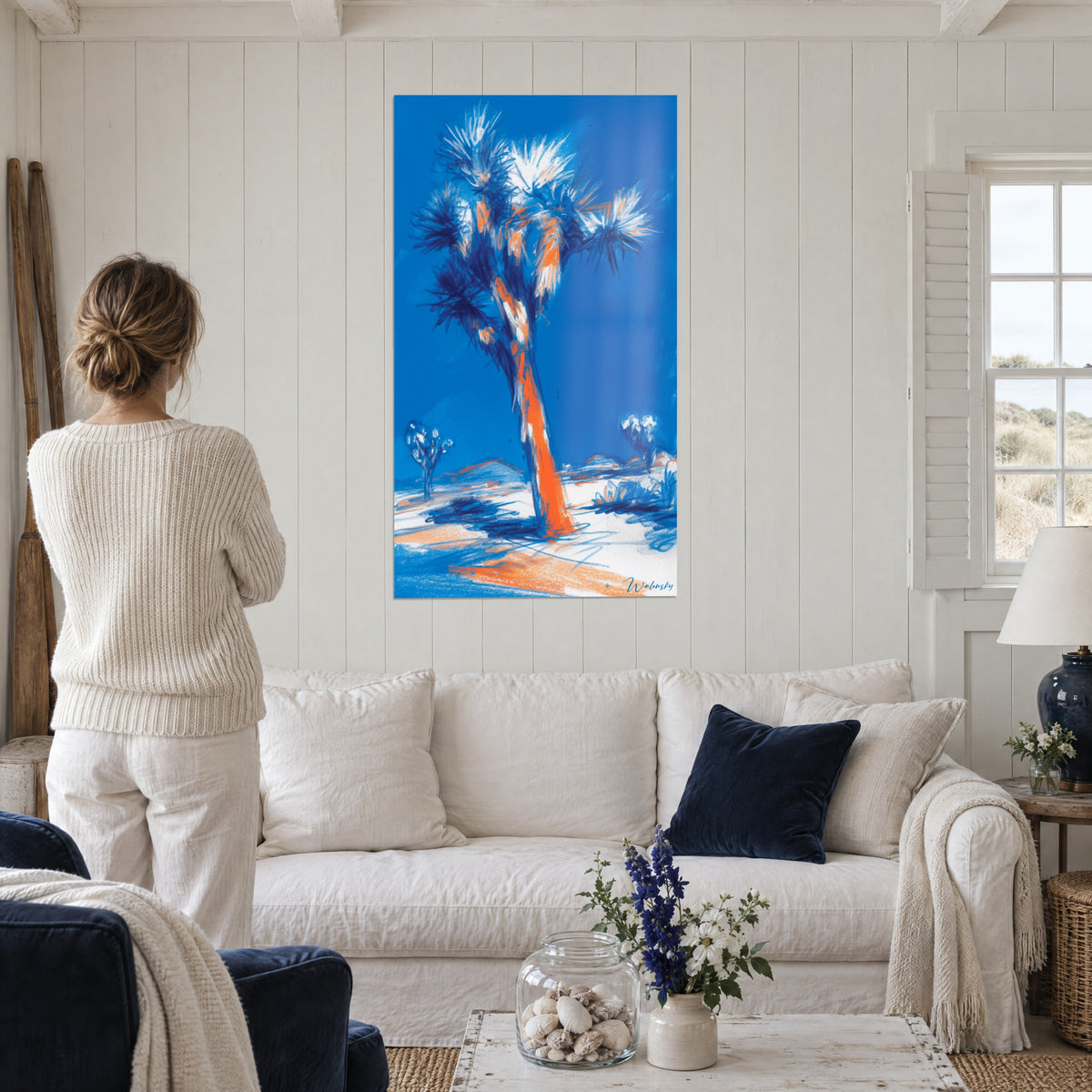 Wall art Joshua tree with white palm fronds, orange trunk against a blue sky, woman seen from behind above a sofa in a bright living room