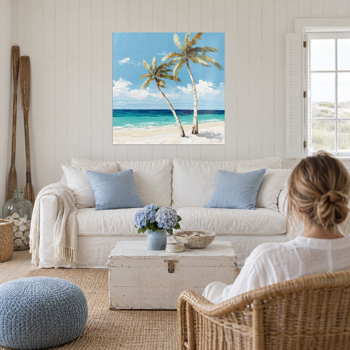 Wall art of two leaning coconut palms with white trunks over turquoise sea, blue sky, woman seen from behind, above a sofa in a bright living room