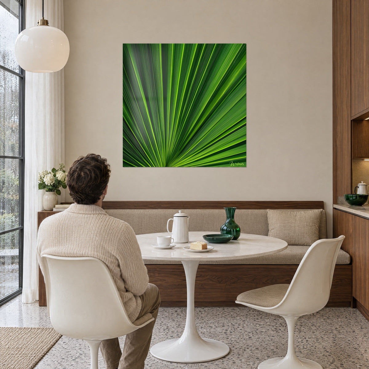 Schilderij van een waaierblad met smaragdgroene en felgele nerven, tegenover een man met zijn rug naar de kijker aan een tafel bij witte stoelen