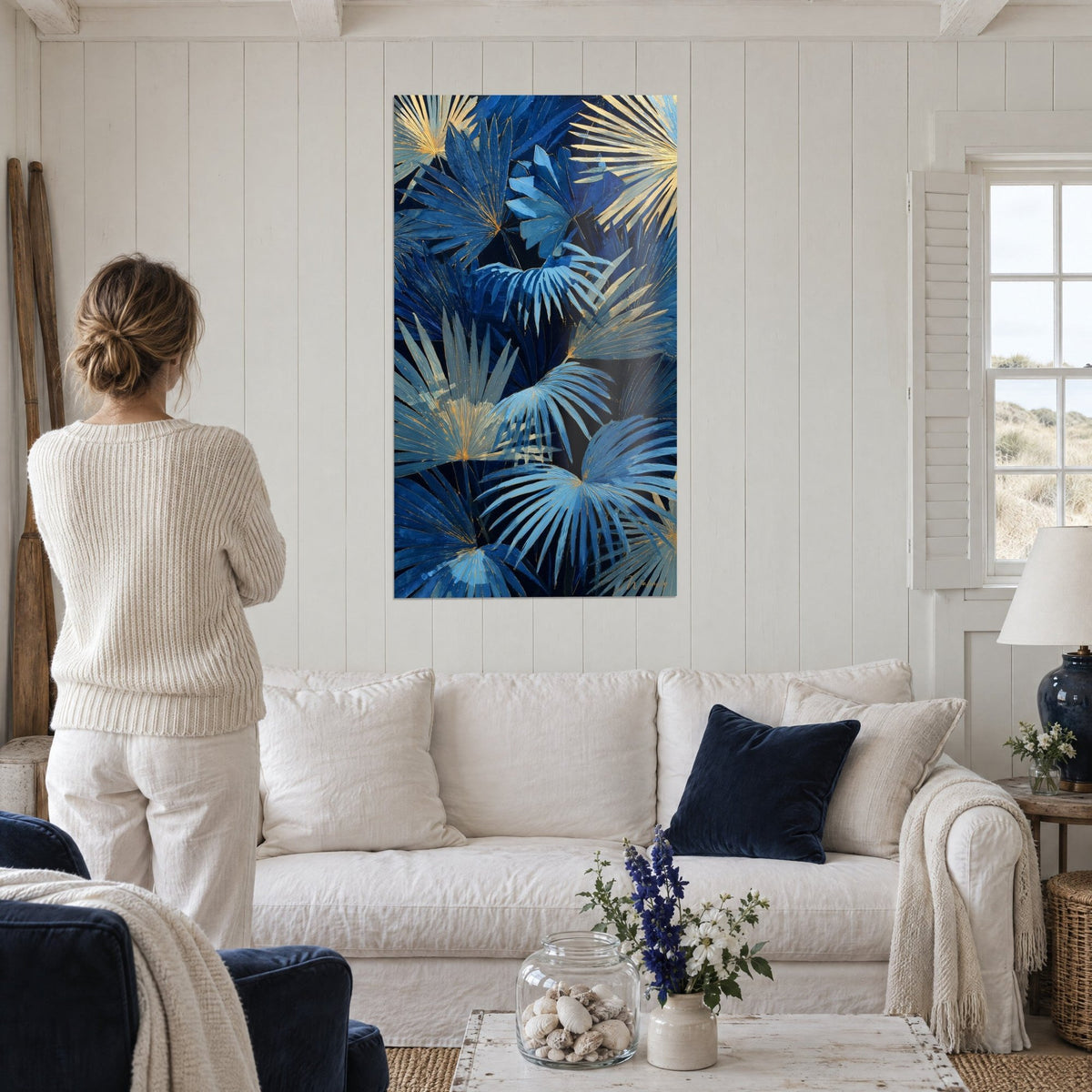 Wall art with navy blue fan palm leaves and gold veins, woman seen from behind, above a sofa in a bright living room