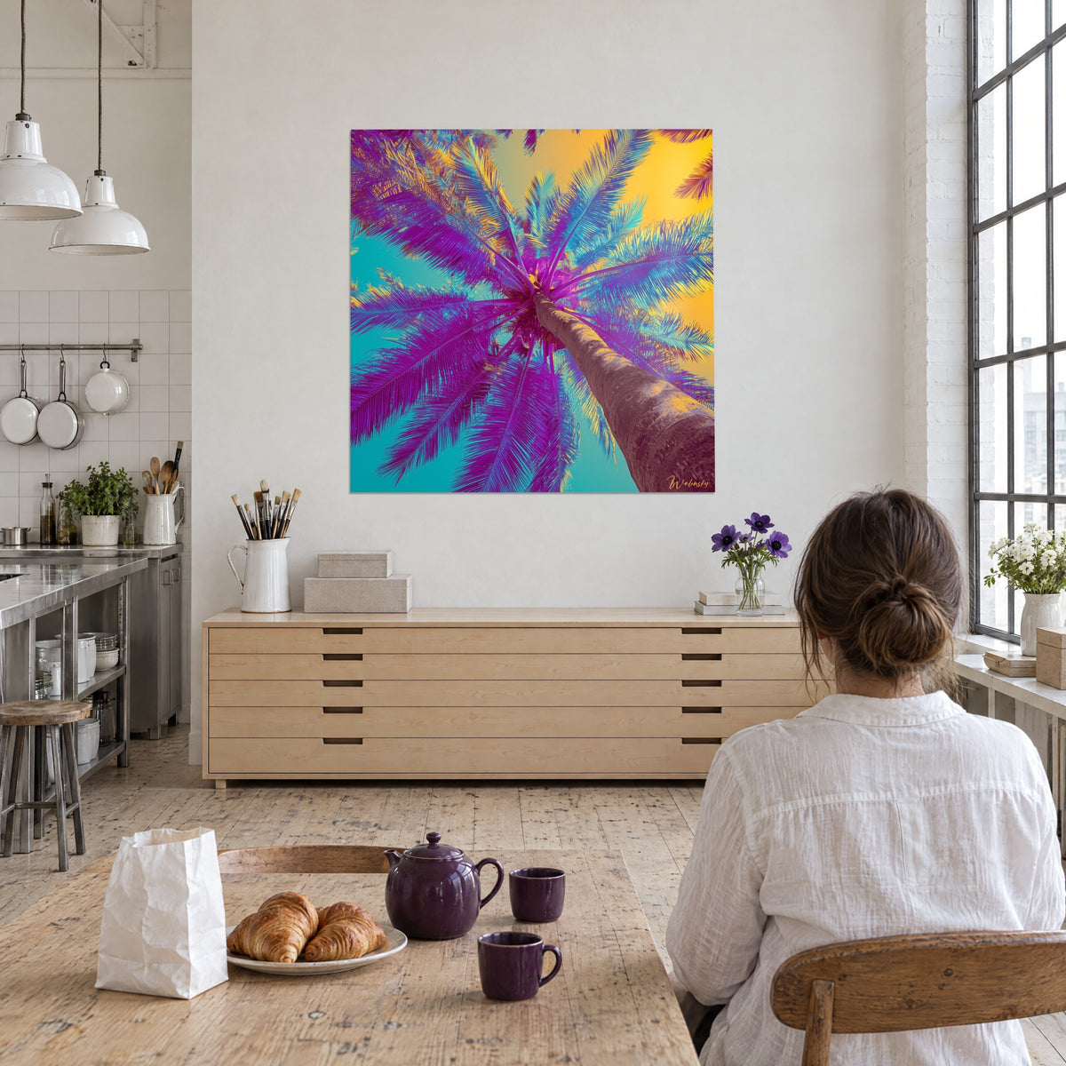 Wall art of a crown of magenta and turquoise palm fronds under a golden sky, viewed by a woman seen from behind in an artist loft