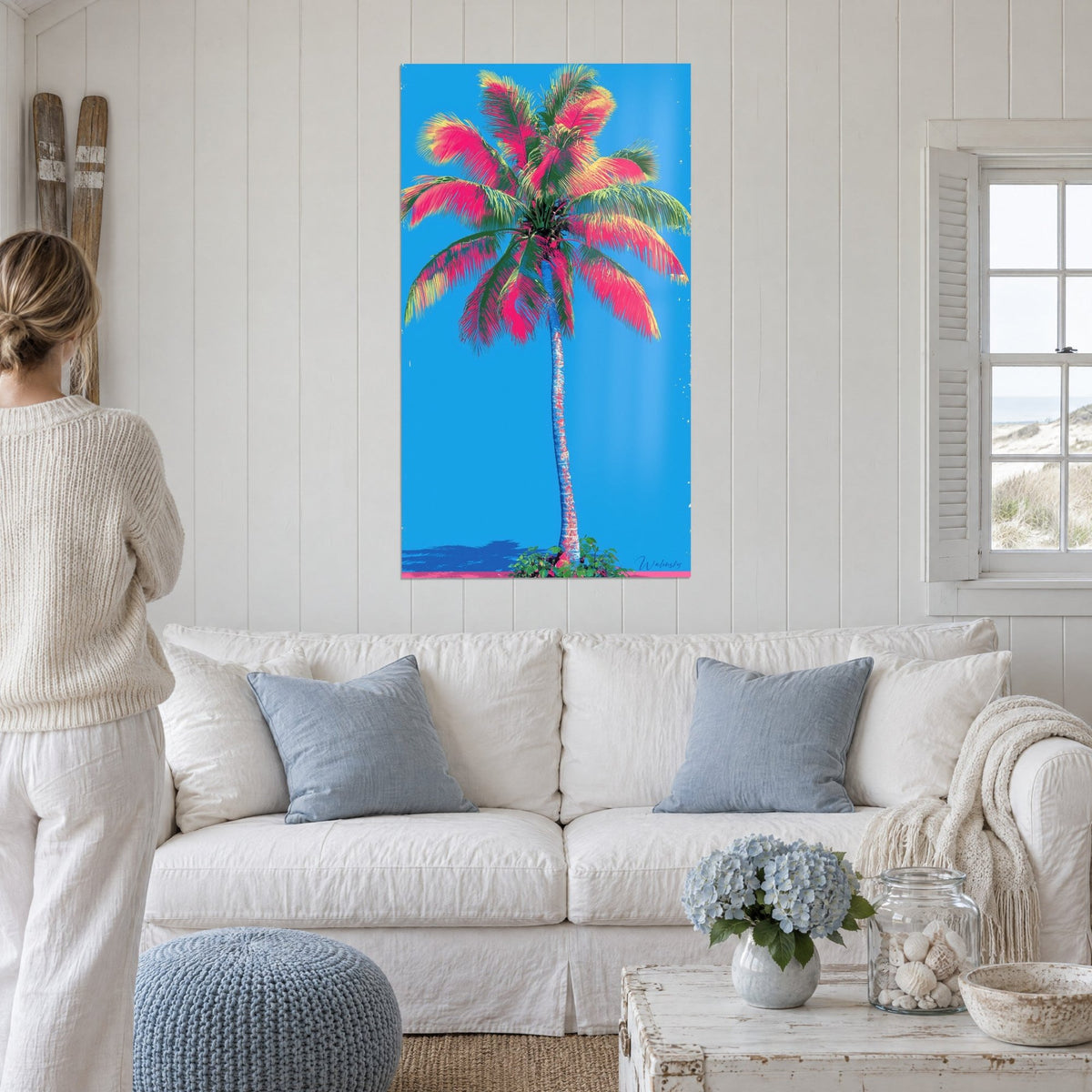 Quadro palma con fronde fucsia e gialle su cielo azzurro, una donna di spalle, sopra un divano in un soggiorno luminoso