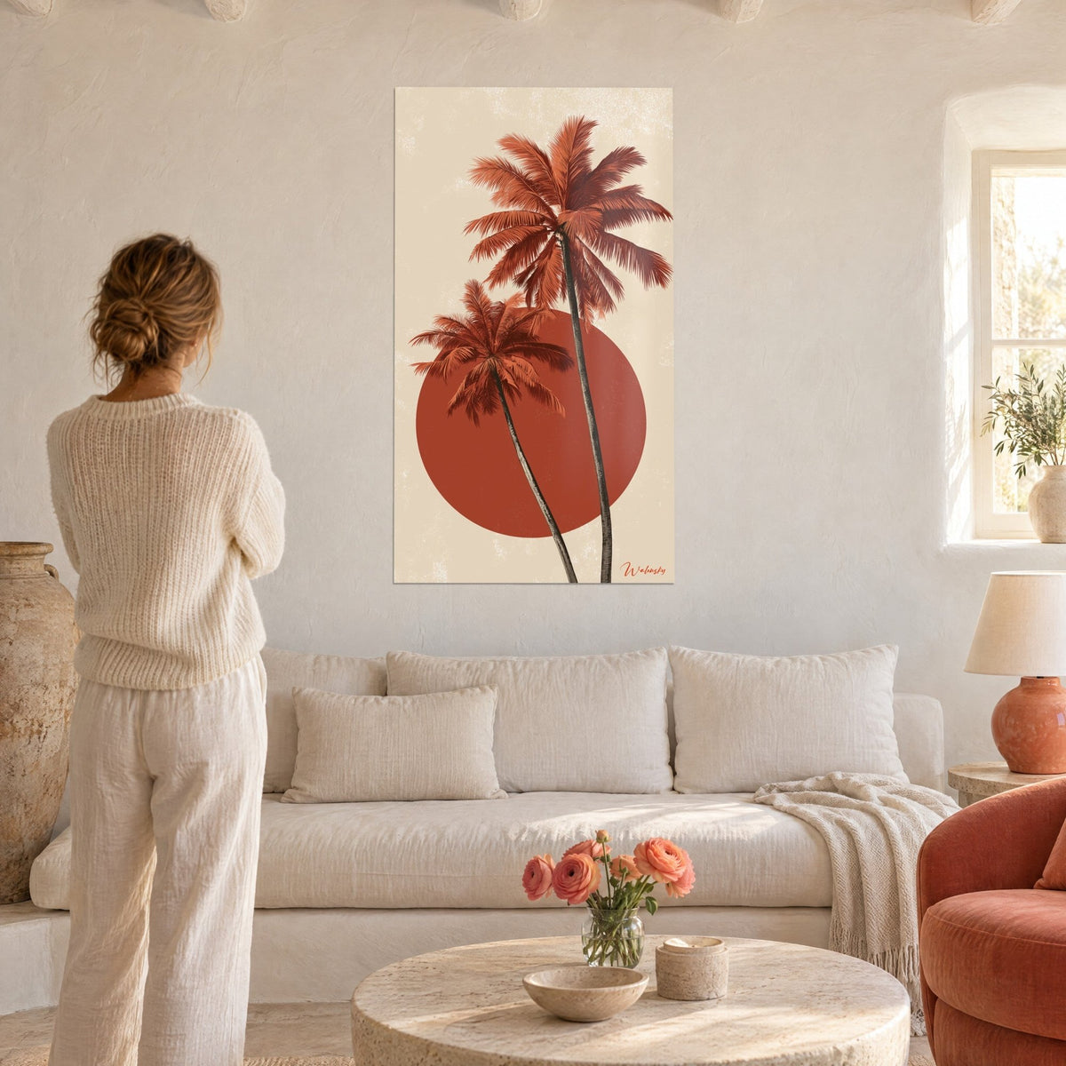 Schilderij terracotta palmbomen die zich buigen voor een rode schijf op een beige achtergrond, vrouw van achteren, boven een bank in een lichte woonkamer