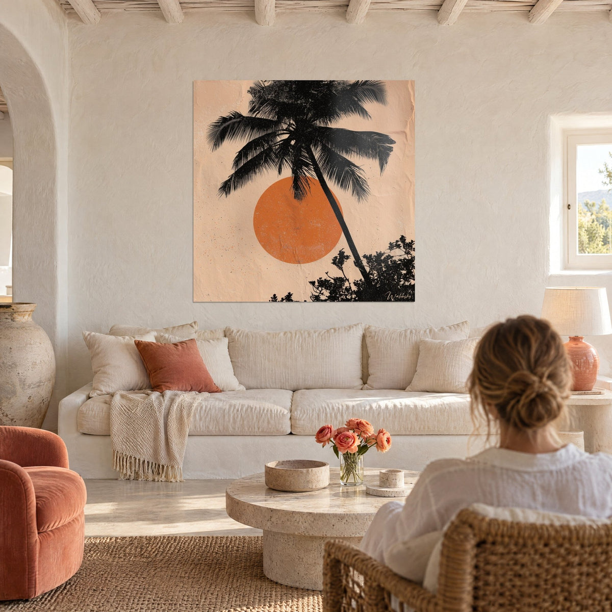 Wall art leaning black palm tree in front of an orange sun on peach background, woman seen from behind above a sofa in a bright living room