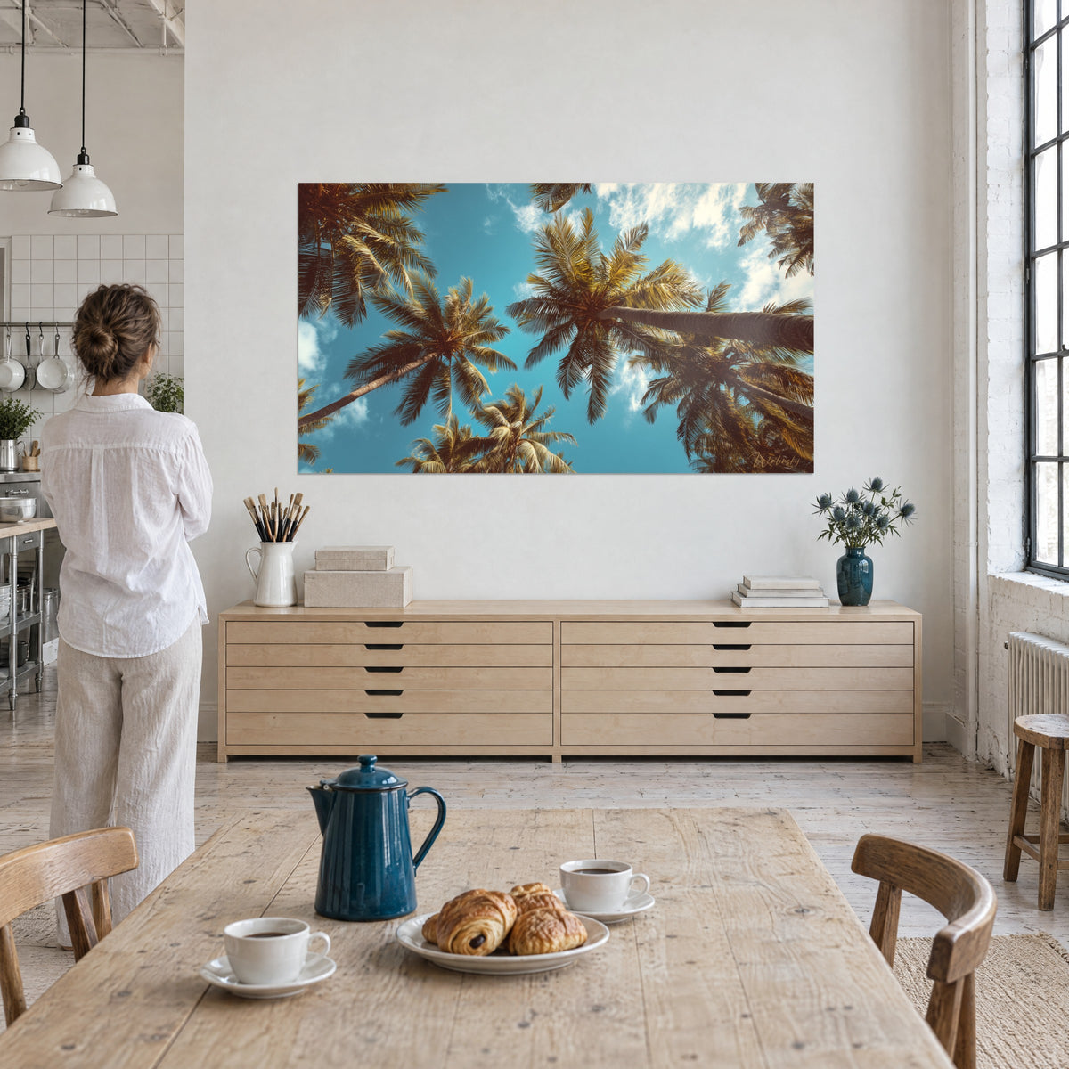 Wall art coconut palms seen from below, slanting brown trunks and cloudy turquoise sky, woman seen from behind in an artist's loft