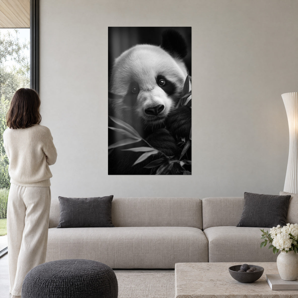 Cuadro de un panda gigante de pelaje blanco crema y hocico negro, hojas de bambú, mujer de espaldas sobre un sofá en un salón luminoso