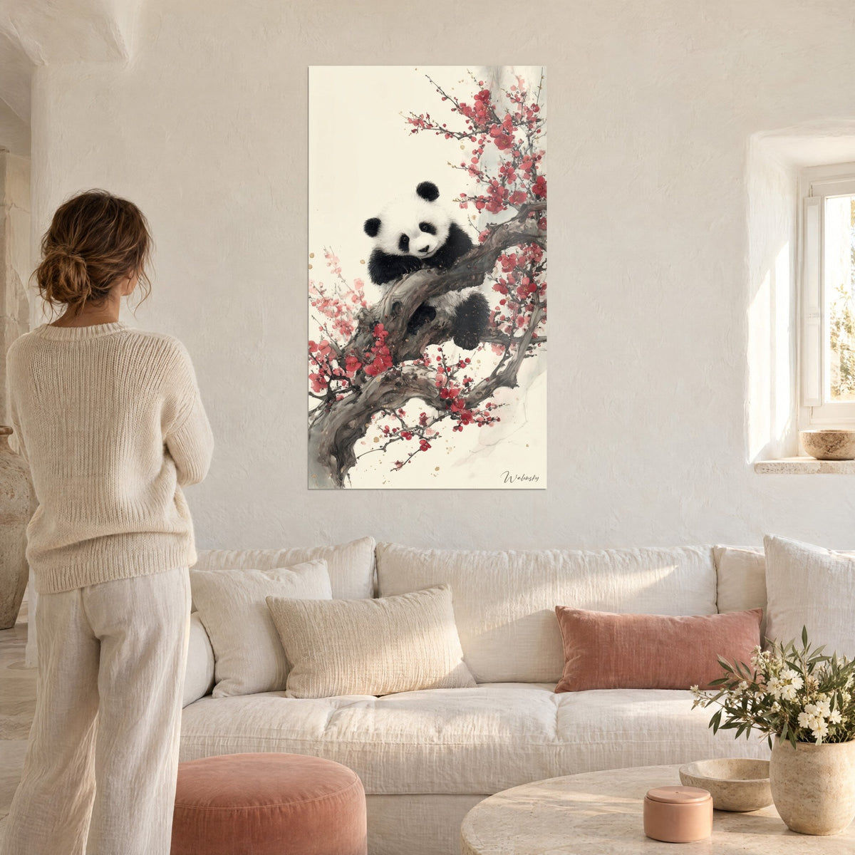 Wall art panda curled up on a branch of a blossoming plum tree with red flowers, cream background, woman seen from behind above a sofa in a bright living room