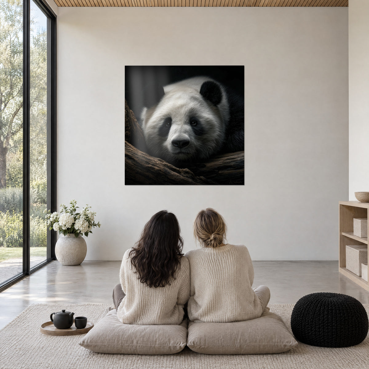 Quadro testa di panda con muso nero e pelliccia crema, ramo marrone scuro, ammirato da due donne sedute di spalle su dei cuscini