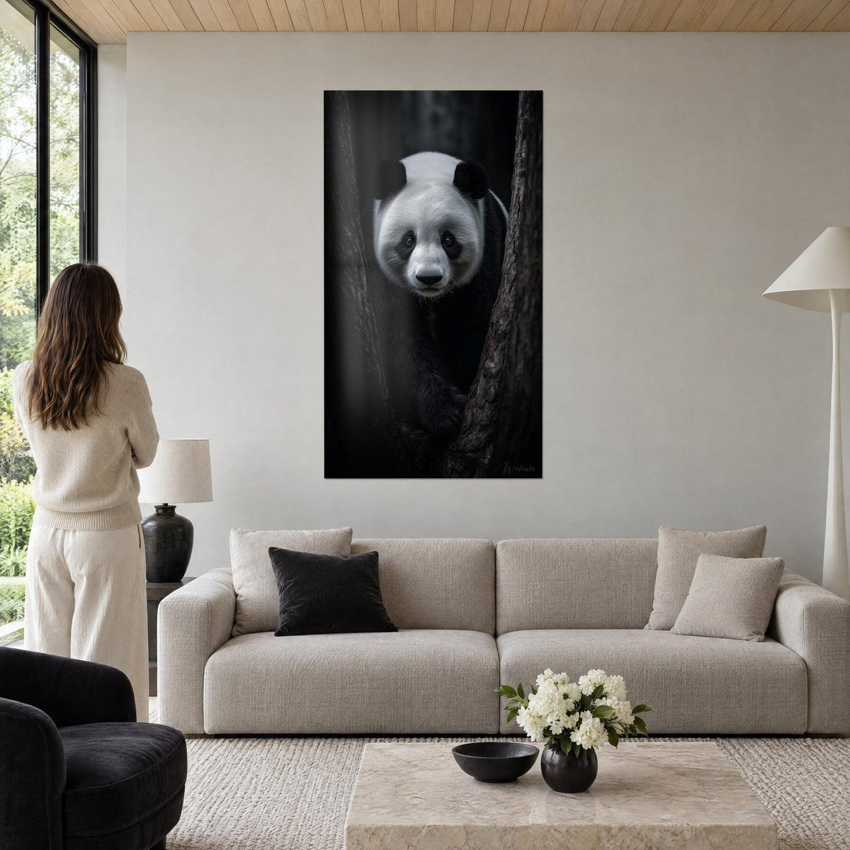 Quadro panda gigante dal pelo avorio e nero corvino, sguardo fisso tra due tronchi, osservato da una donna, sopra un divano in un soggiorno luminoso