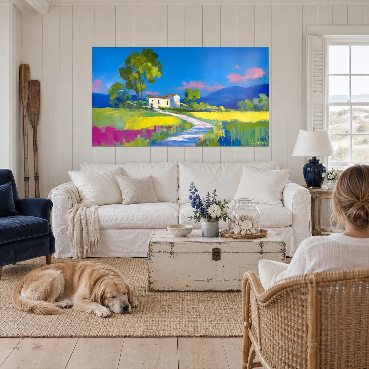 Schilderij witte boerderij met bruin dak onder een intens blauwe lucht, gele en roze weide, boven een bank in een lichte woonkamer