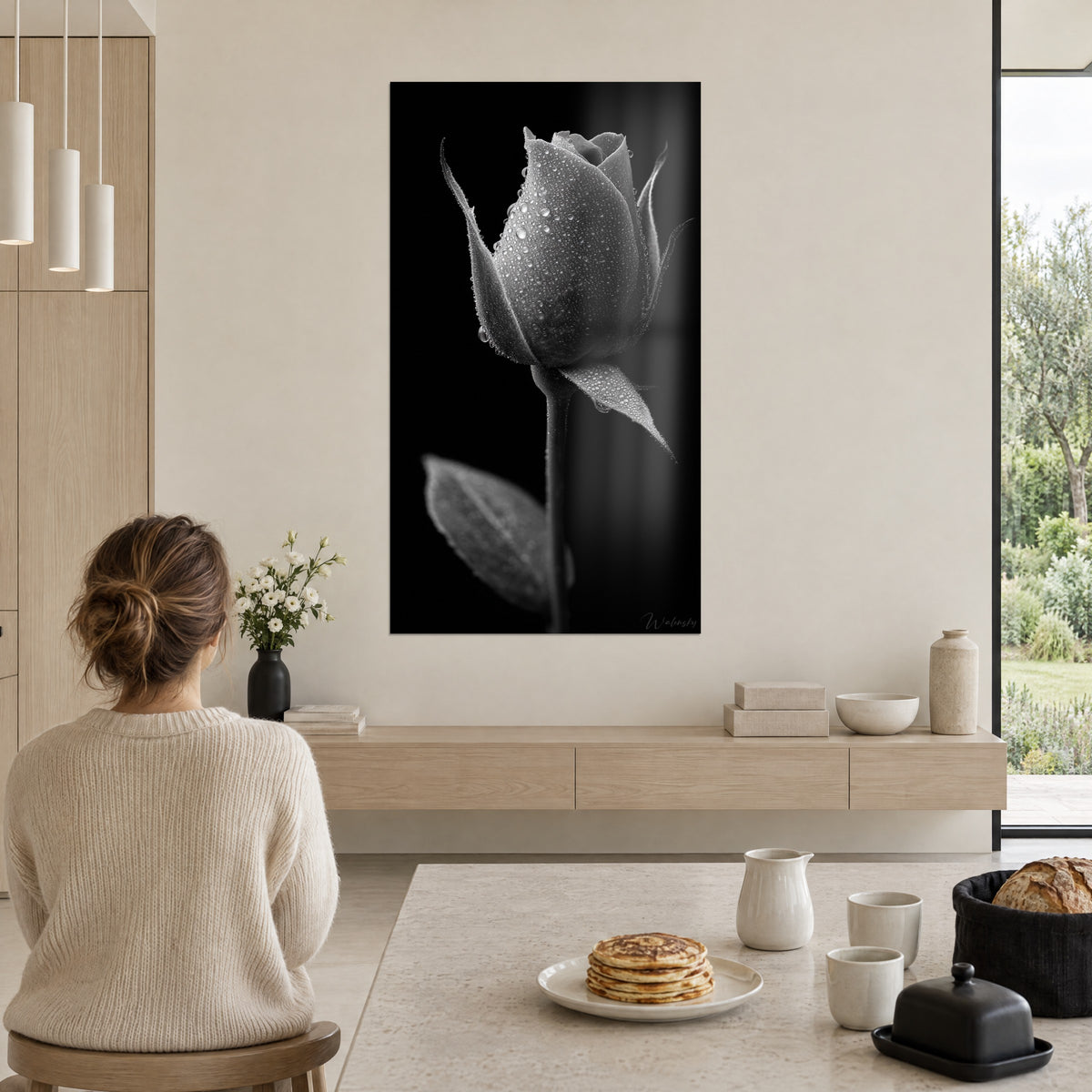 Cuadro rosebud plateado salpicado de gotas, fondo negro y hoja gris perla, mujer de espaldas sentada a la mesa, pared blanca, ventanal abierto