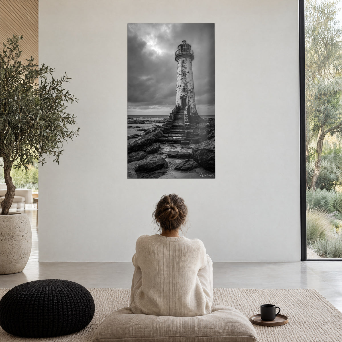 Schilderij wachter van afbladderend wit steen tegenover de grijze zee, vrouw van achteren zittend in een crèmekleurige trui, zwarte poef, raam tot de vloer met uitzicht op het groen