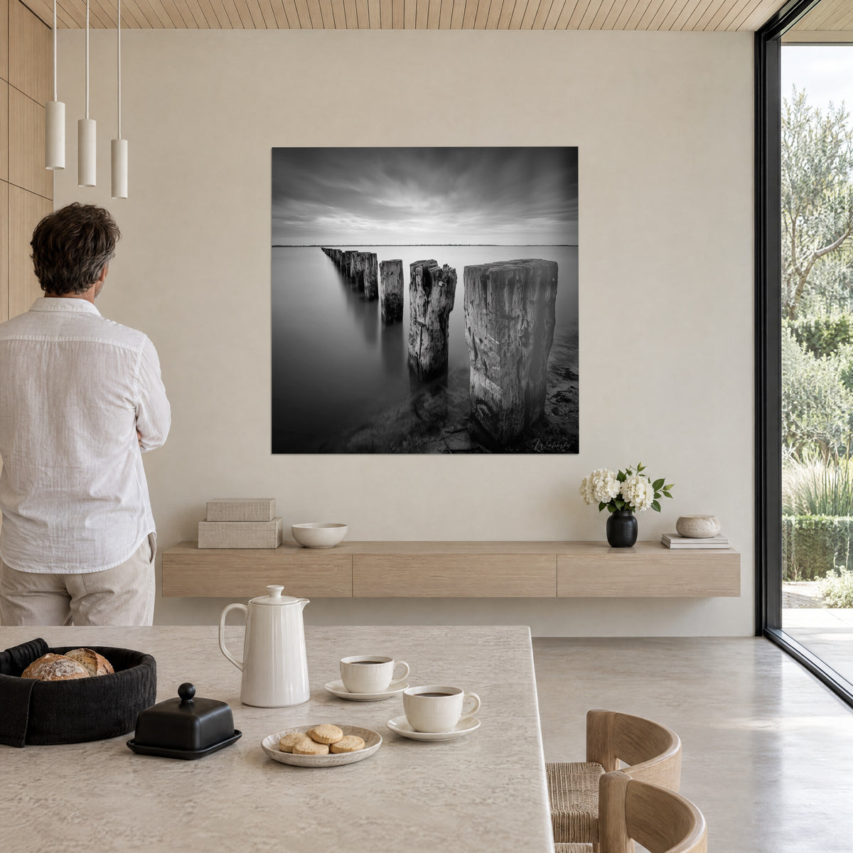 Tableau estacade de troncs usés dans une mer d'huile, gris argenté et noir velouté, ciel de longue pose, devant un homme de dos dans un séjour