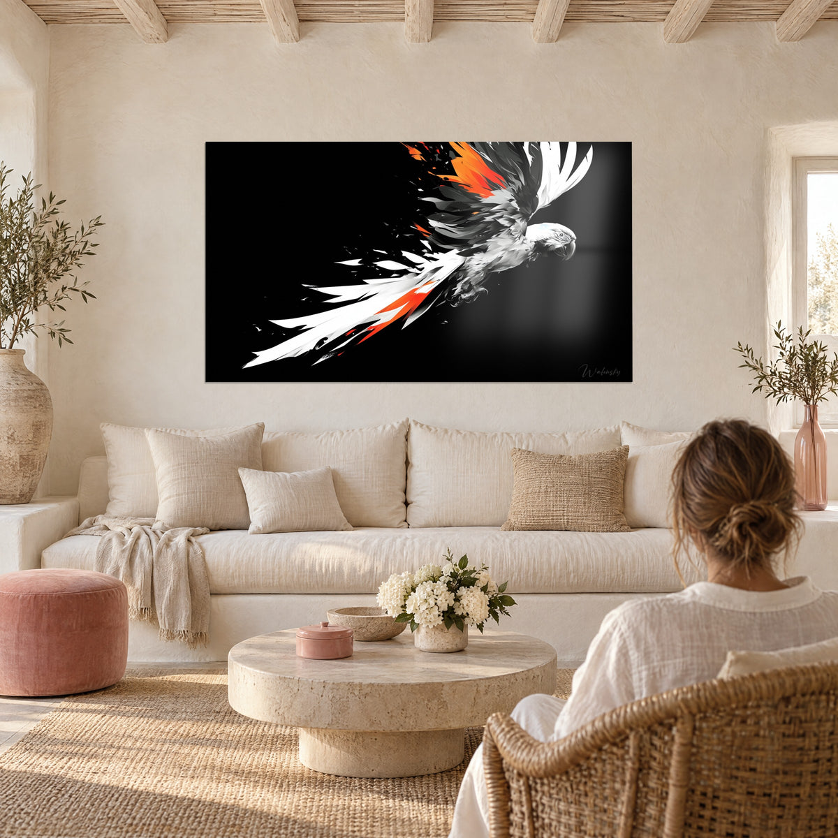 Wall art of a white parrot with vivid orange feathers in full flight against a black background, hung above a sofa in a bright living room with a woman seen from behind