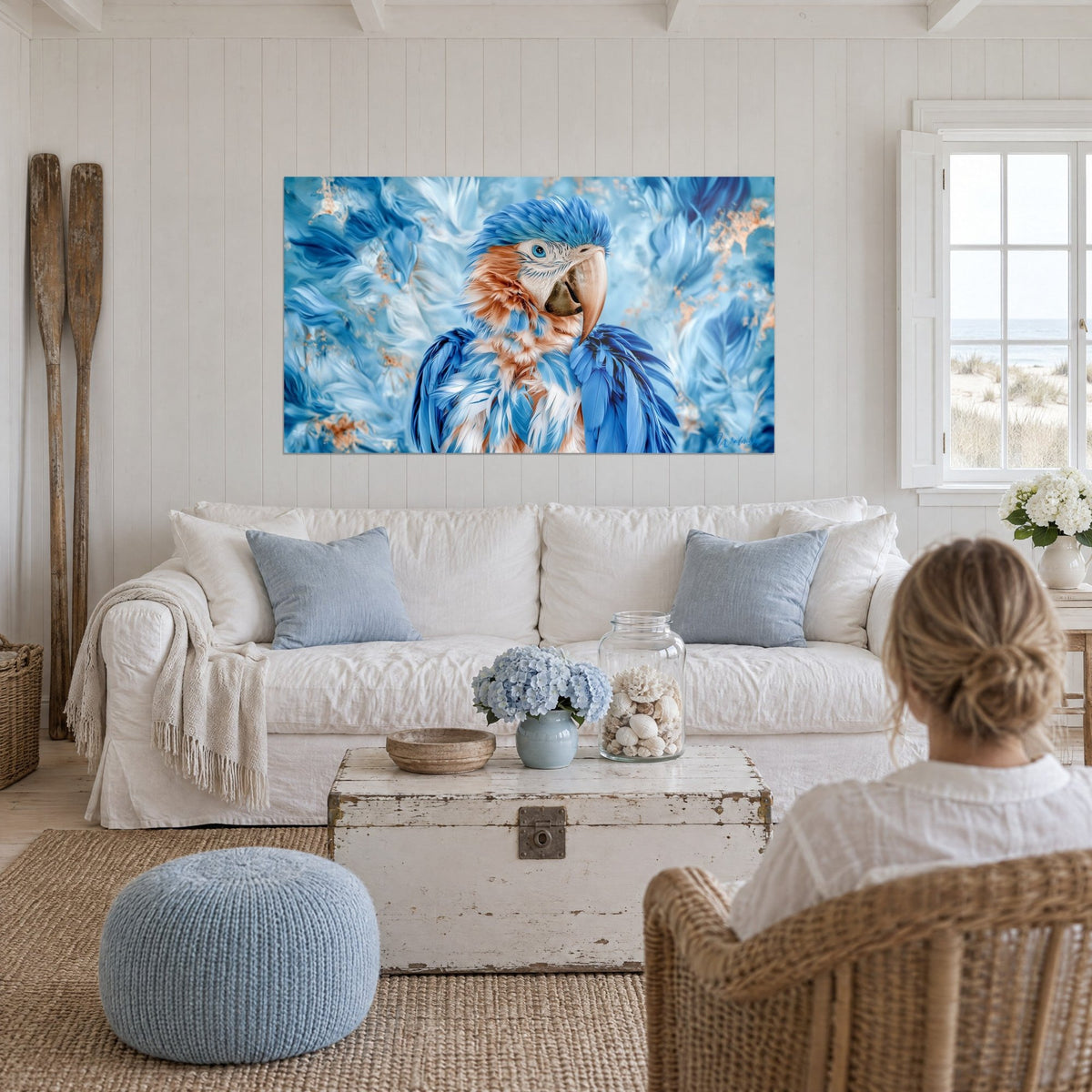 Gemälde Ara in Blau mit pfirsichfarbenem Schnabel und seidigem Gefieder, eisblauer Hintergrund, Frau von hinten, über einem Sofa in einem hellen Wohnzimmer