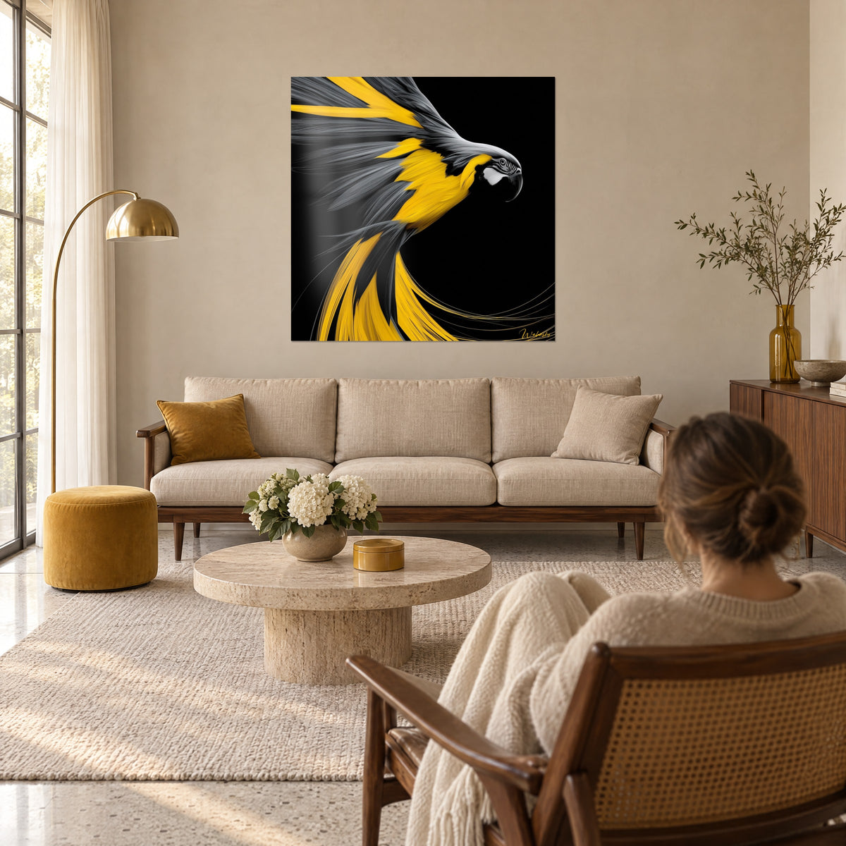 Wall art of a sunny yellow macaw with grey wings spread wide on a black background, gazed at by a woman, above a sofa in a bright living room