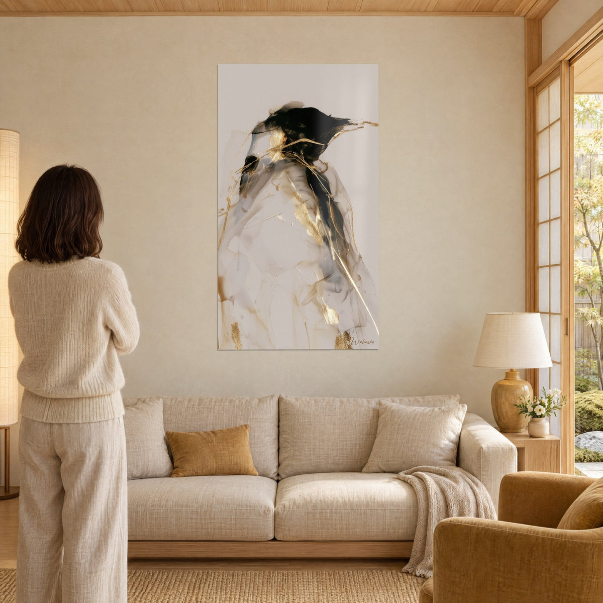 Tableau manchot au plumage noir et ivoire, filaments d'or sur fond beige, regardé par une femme de dos, au-dessus d'un canapé dans un salon lumineux
