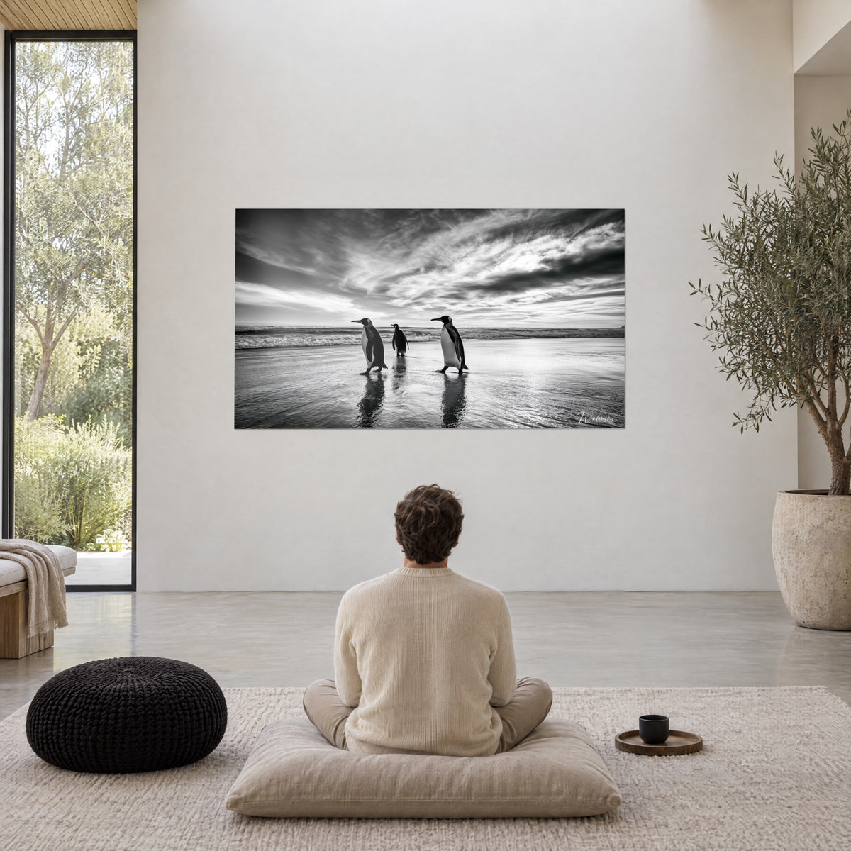 Tableau marche de trois pingouins sur la grève en monochrome, houle grise et ciel dramatique, un homme assis sur un coussin le contemple