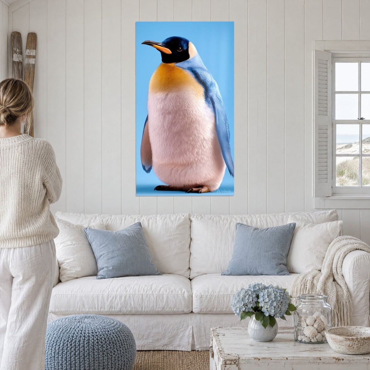 Tableau manchot au plastron doré et ailes bleues sur fond ciel, accroché au-dessus d'un canapé dans un salon lumineux