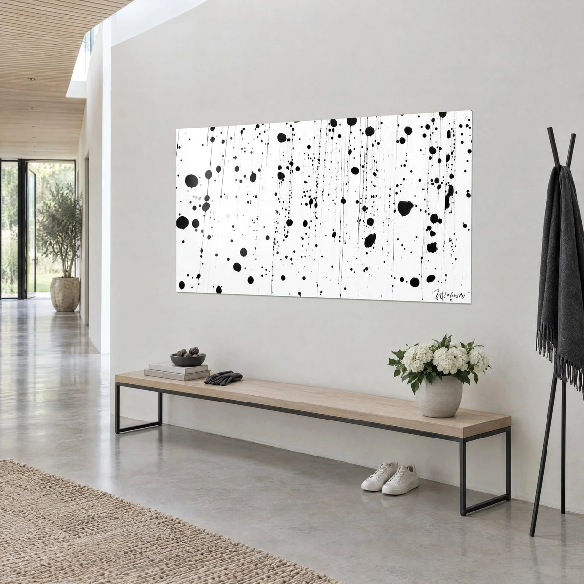 Quadro gocce nere e tratti filiformi su bianco puro, appeso a una parete grigia, panca in legno chiaro e fiori bianchi
