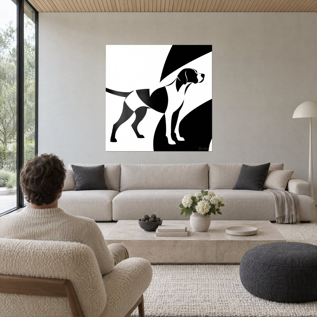 Wall art of a stylised hunting dog in flat black shapes on a white background, tail stretched out, man seen from behind, above a sofa in a bright living room