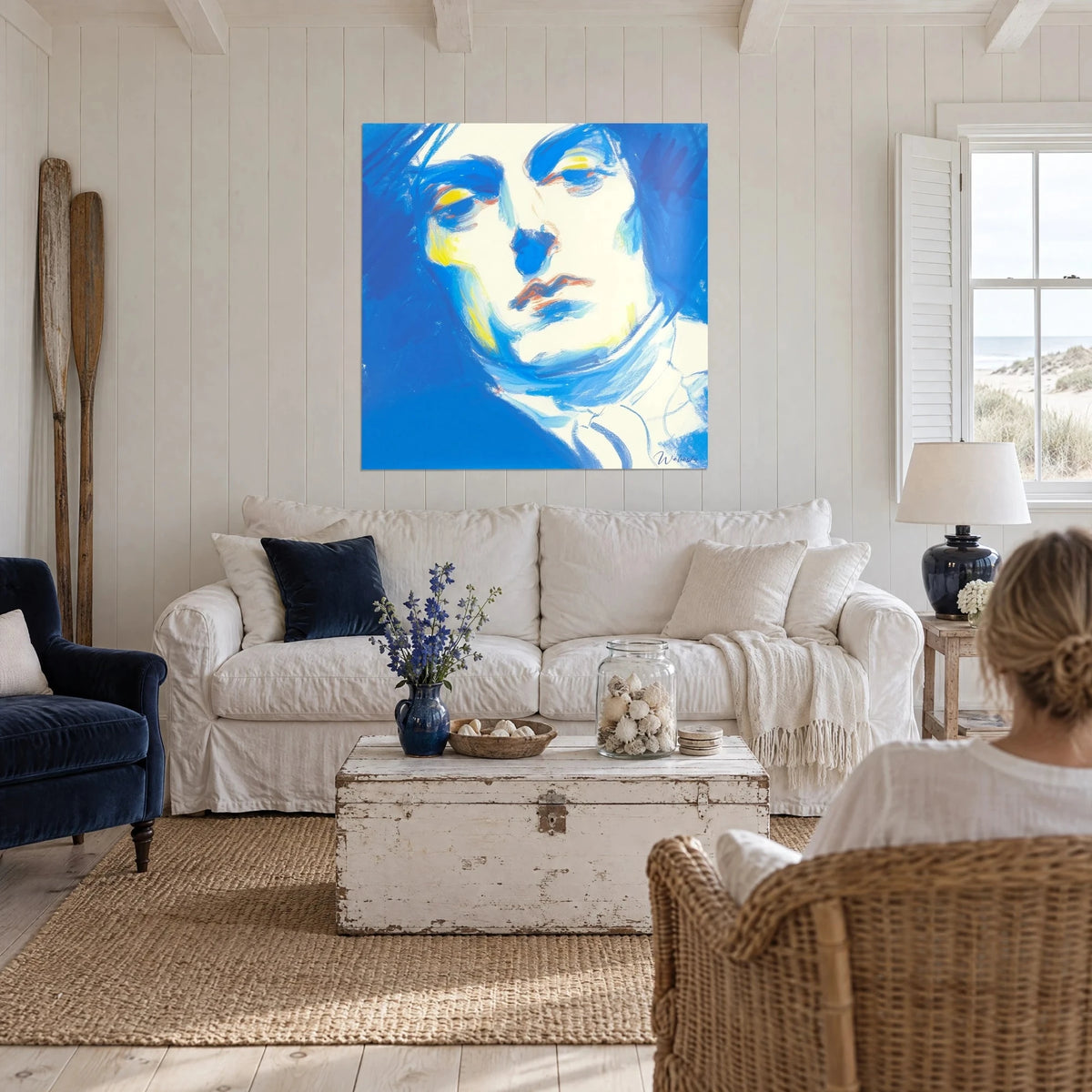 Wall art of a solemn face with cobalt blue shadows and lemon yellow cheeks, woman seen from behind, above a sofa in a bright living room
