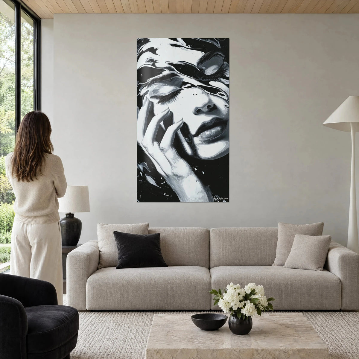 Wall art woman's face with closed eyelids, hand with long nails resting on the cheek, black and white, above a sofa in a bright living room