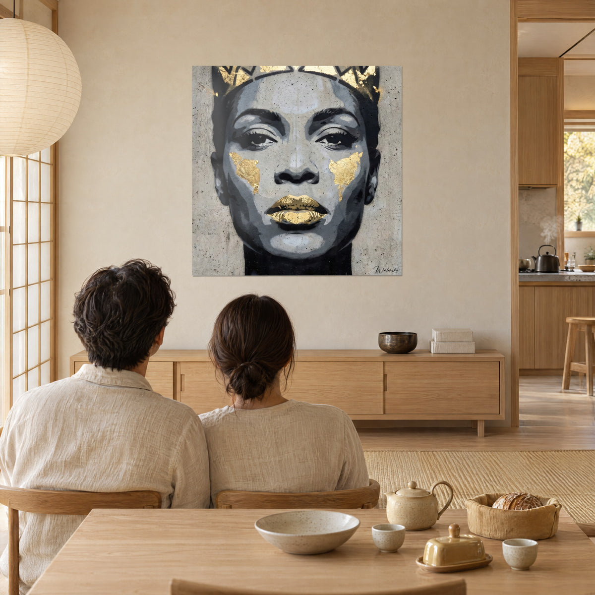Quadro effigie a stencil con zigomi dorati, tonalità di grigio e oro, di fronte a una coppia seduta di spalle a un tavolo in legno chiaro