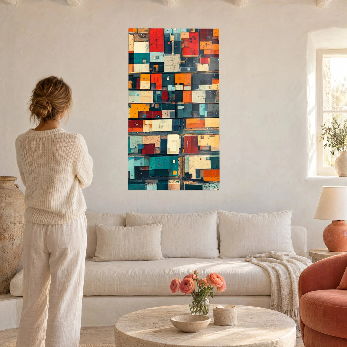 Wall art of patchwork rooftops seen from above, vermilion red and turquoise, woman seen from behind, above a sofa in a bright living room