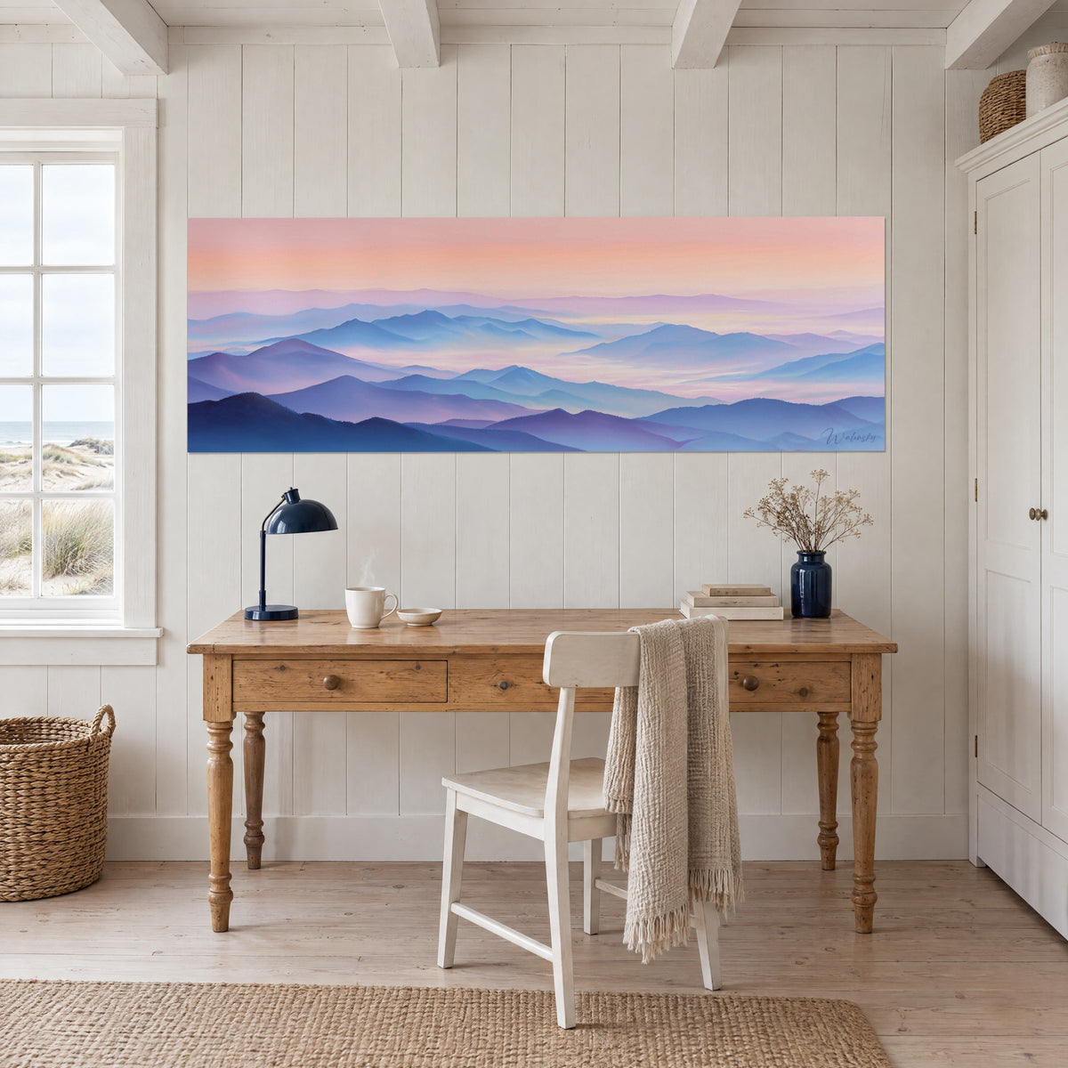 Wall art of an indigo blue and pale pink mountain landscape, light wood desk with a navy blue lamp, by the seaside