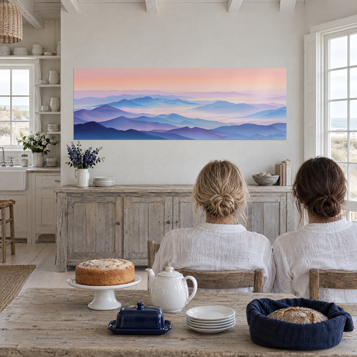 Wall art of misty valleys in steel blue and mauve, apricot sky, two people seen from behind seated at a table beside a cake, beach house