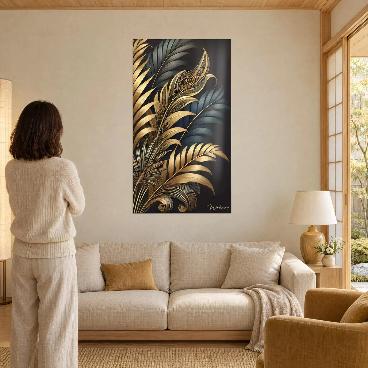 Wall art with gilded, ribbed palm fronds on a black background, blue-grey leaves in the back, woman seen from behind, above a sofa in a bright living room