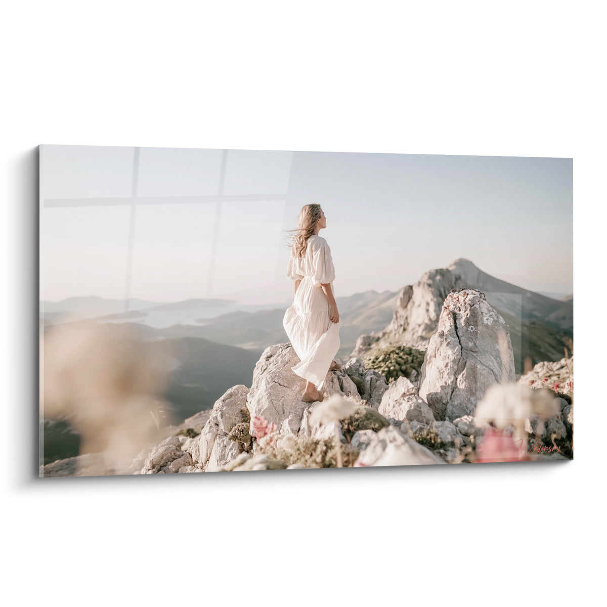 Randonneuse en robe blanche debout sur un rocher gris face aux montagnes bleu pastel