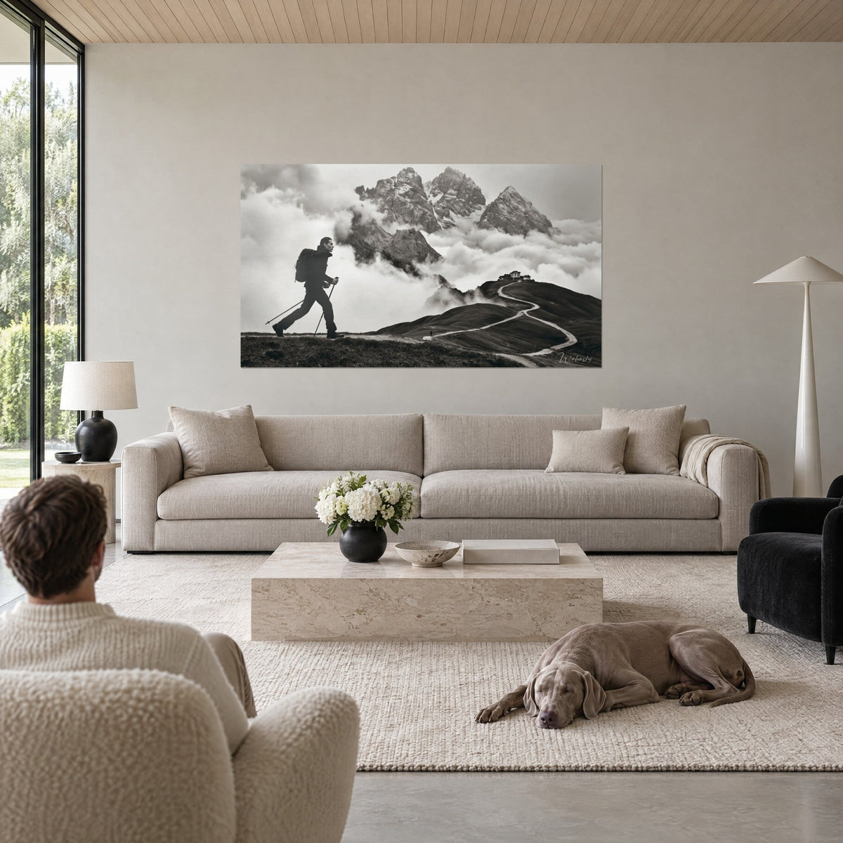 Tableau marcheur à bâtons en silhouette noire, chemin blanc sinueux et cimes grises dans la brume, au-dessus d'un canapé dans un salon lumineux