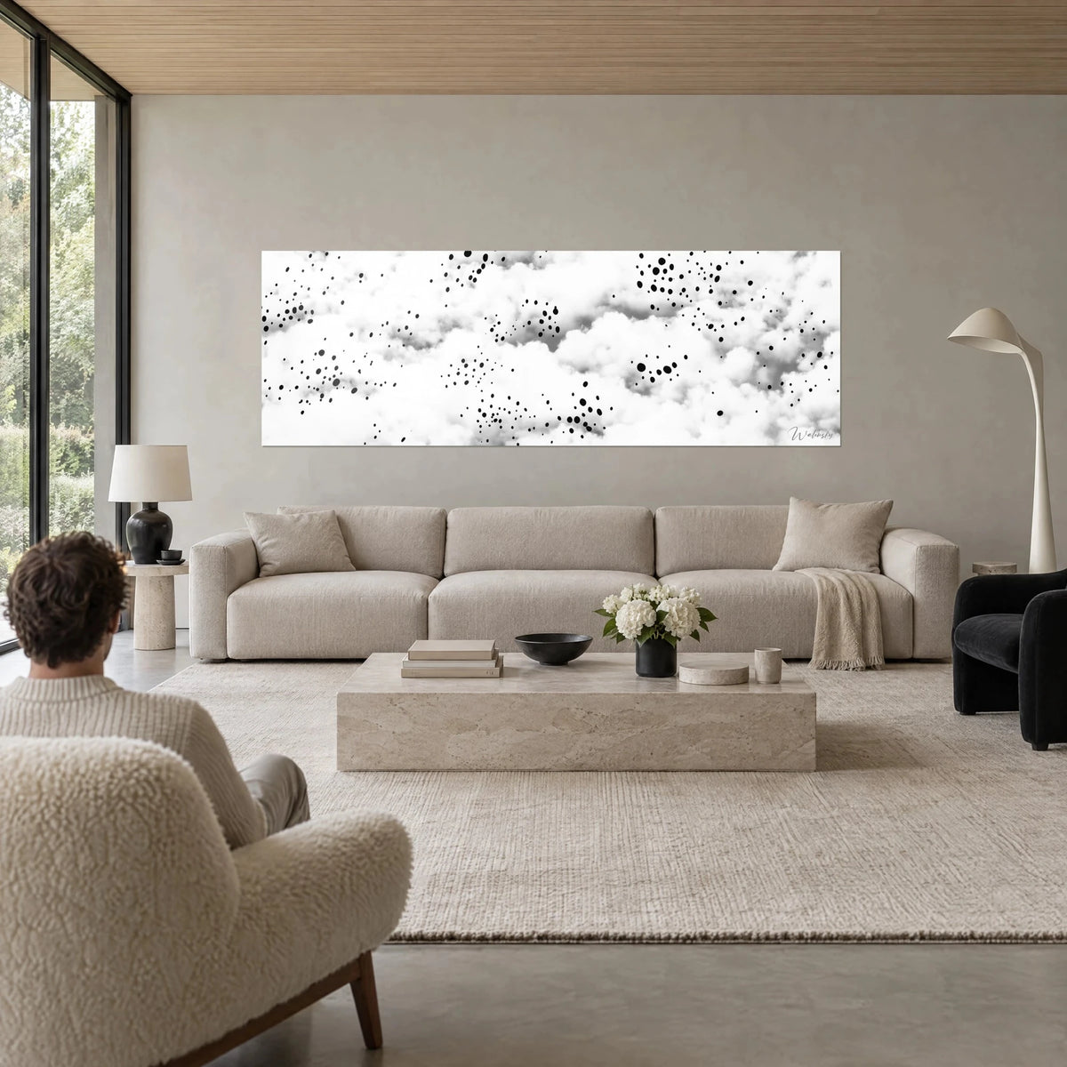 Wall art of white clouds speckled with black ink drops, pearl grey sky, person seen from behind, above a sofa in a bright living room