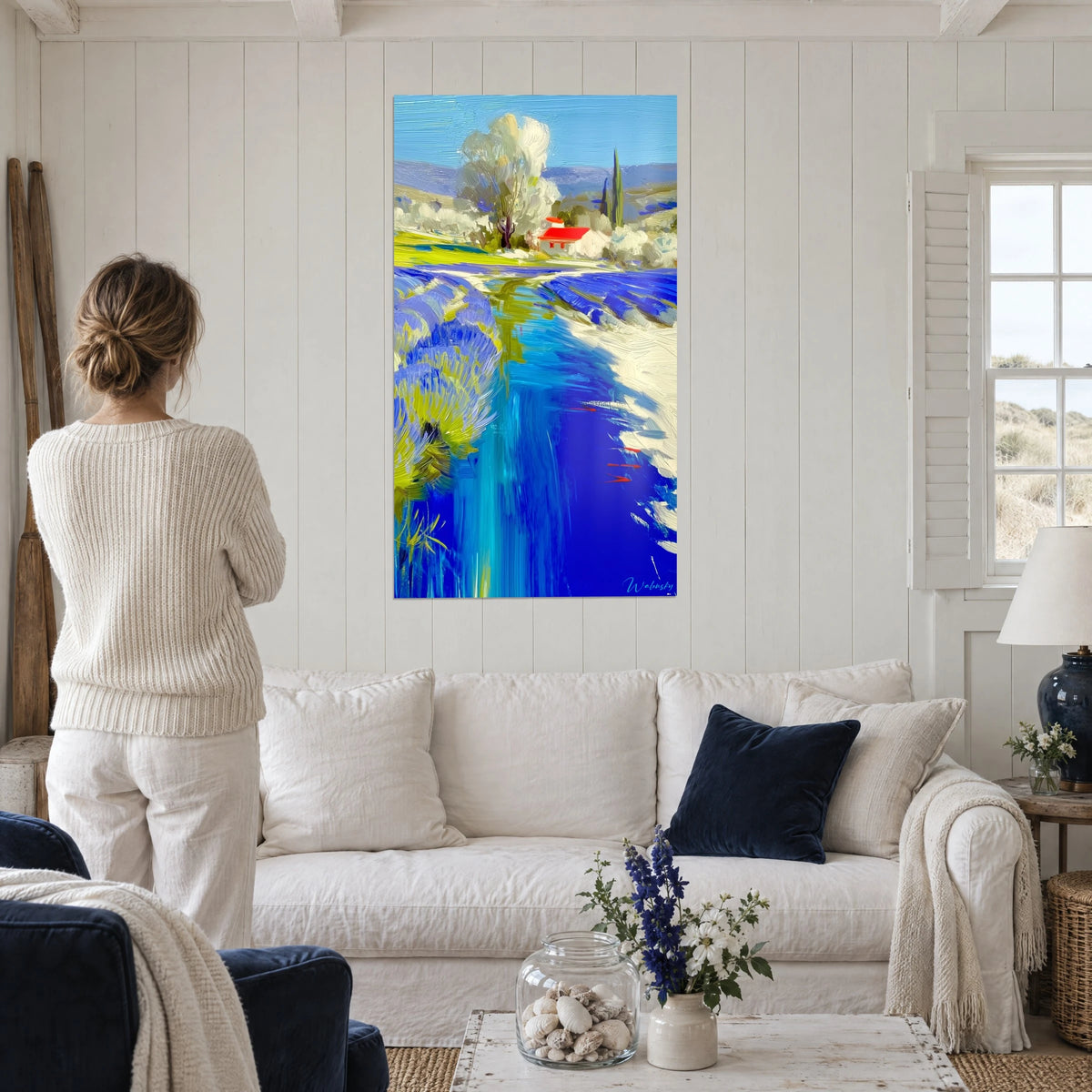 Quadro ruscello blu cobalto tra file di lavanda, tetto rosso e albero chiaro, sopra un divano in un soggiorno luminoso