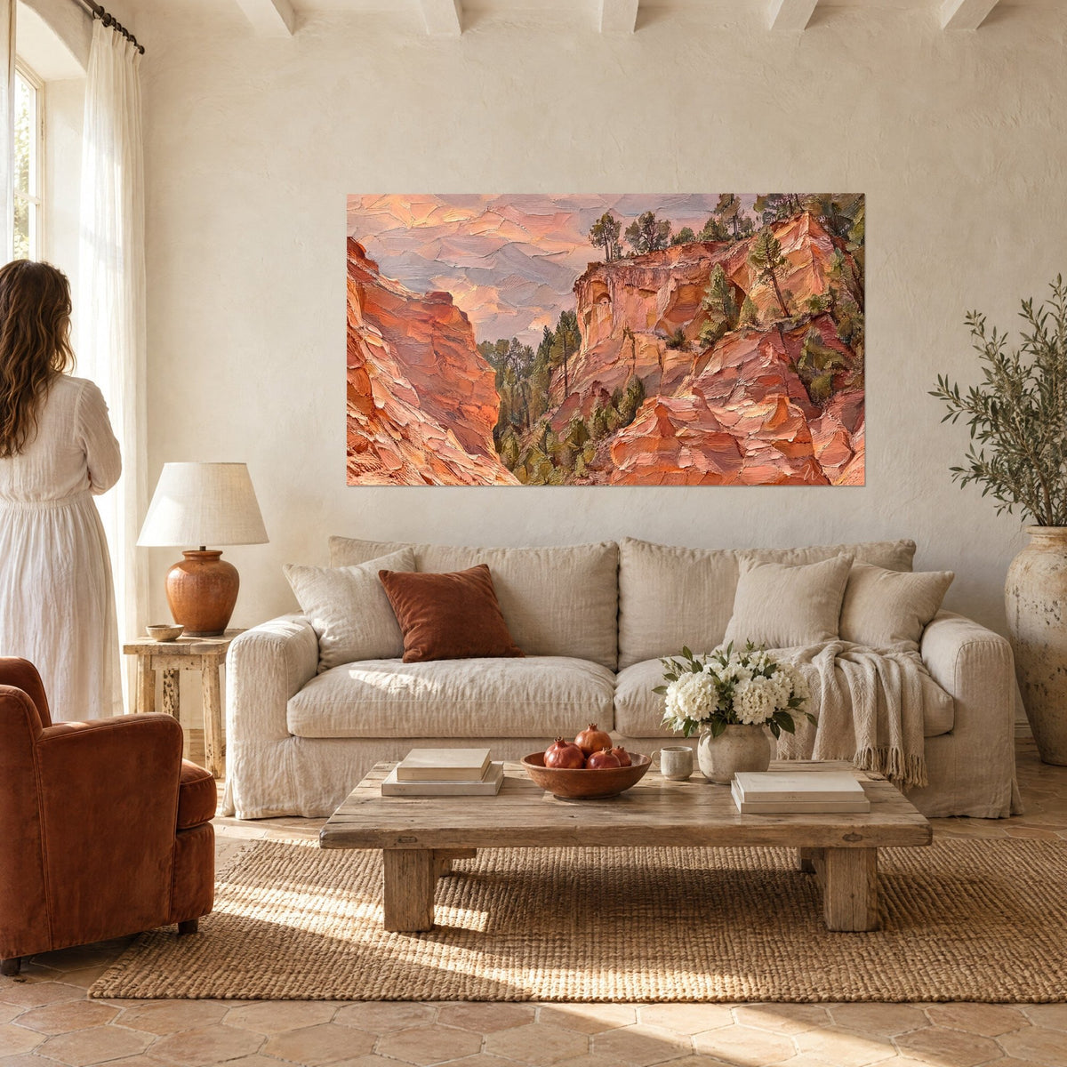 Wall art of red ochre cliffs and green pines under a pink sky, a woman seen from behind above a sofa in a bright living room
