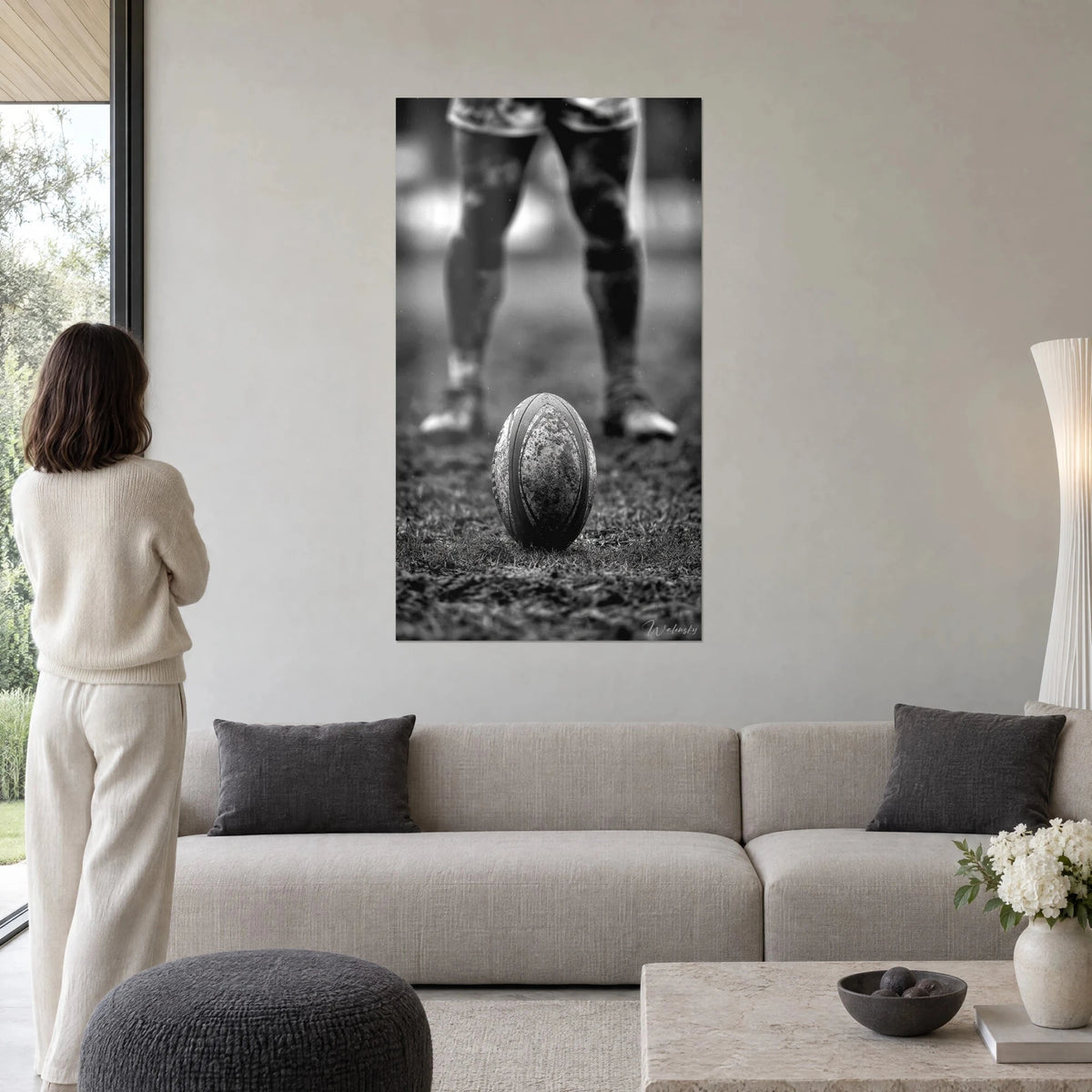 Wall art of a mud-covered rugby ball on a waterlogged pitch, a player's legs in grey and black, a woman seen from behind above a sofa in a bright living room