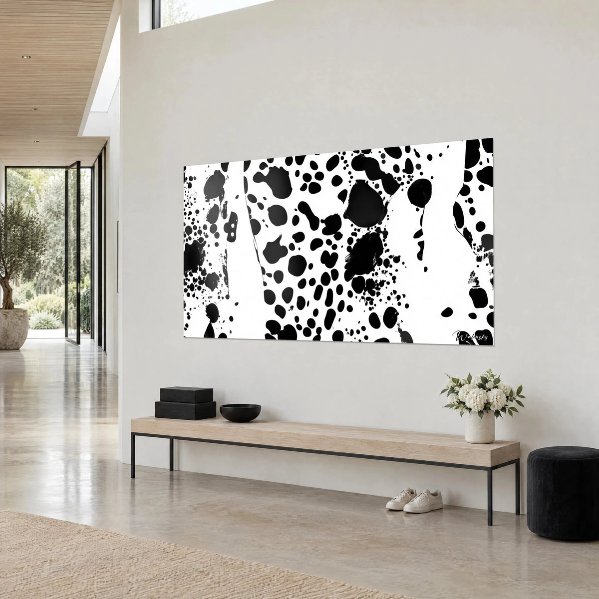 Wall art with black speckles in an irregular rhythm on pure white, hung above a light wood bench and a black pouf