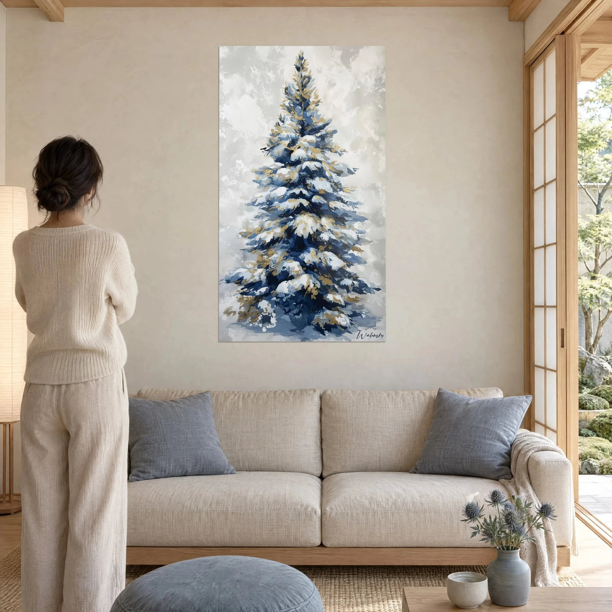 Wall art snowy fir tree with navy blue branches and gold glitter accents, woman seen from behind, above a sofa in a bright living room