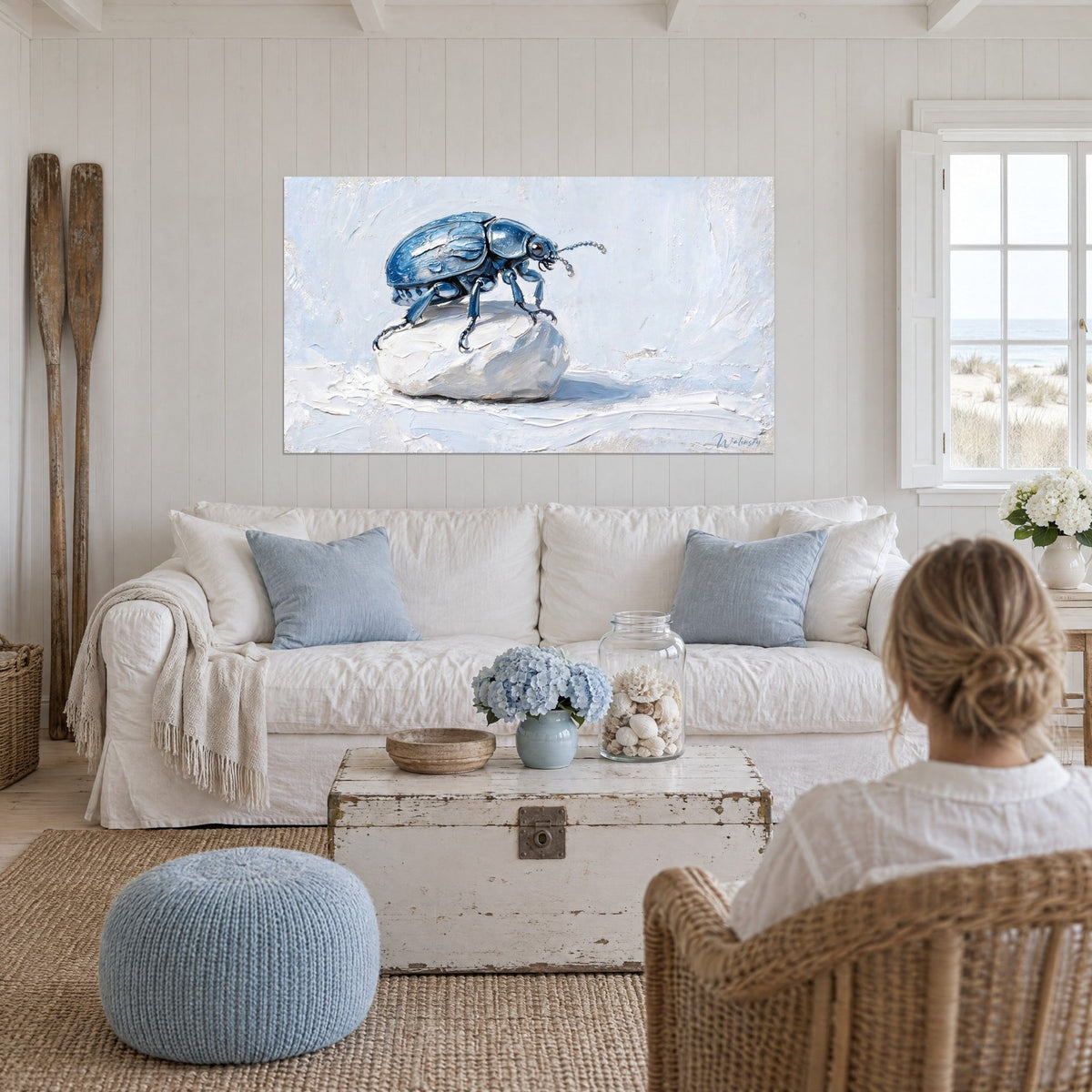 Tableau scarabé aux élytres bleu acier perché sur un galet blanc, fond bleu pâle, au-dessus d'un canapé dans un salon lumineux