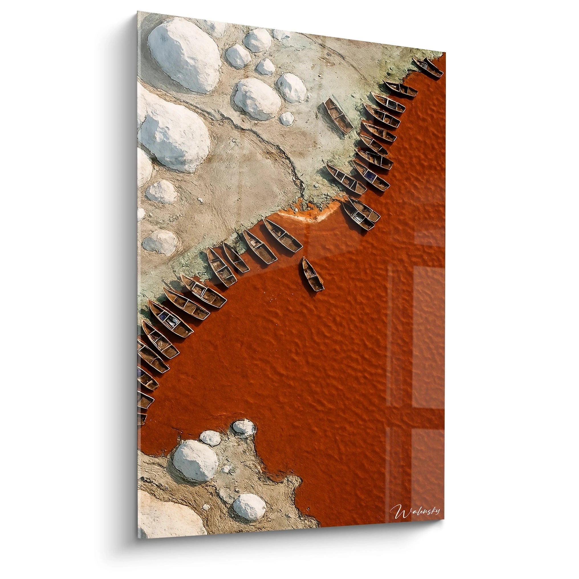 Un tableau vue aérienne montrant des barques en bois alignées entre une eau rouge-orangée et une rive beige. Des formations blanches circulaires parsèment le rivage, créant un contraste saisissant avec la couleur vive de l'eau.

