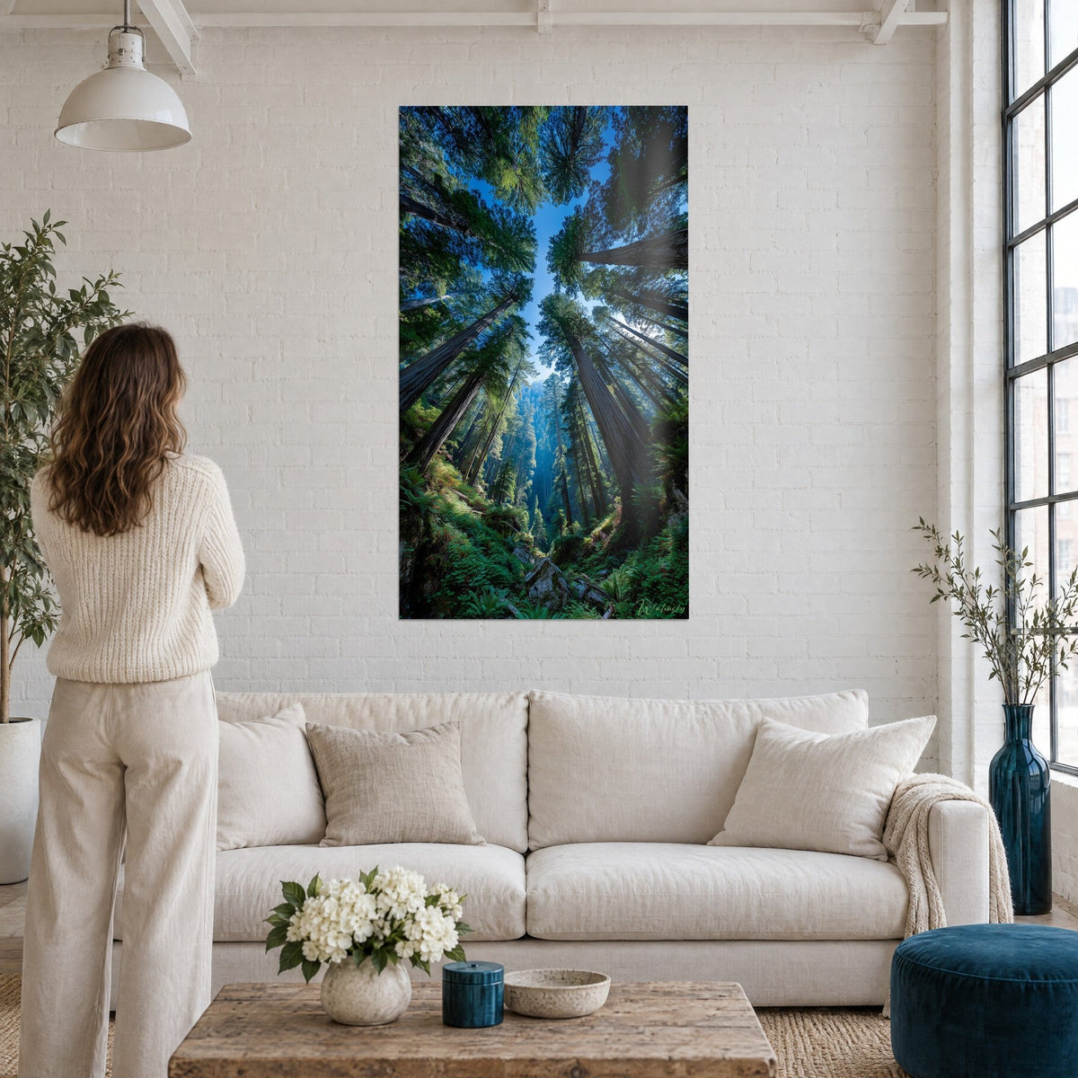 Schilderij reusachtige sequoia's van onderaf gezien, roodbruine stammen, groene kruinen en blauwe lucht, vrouw van achteren, boven een bank in een lichte woonkamer