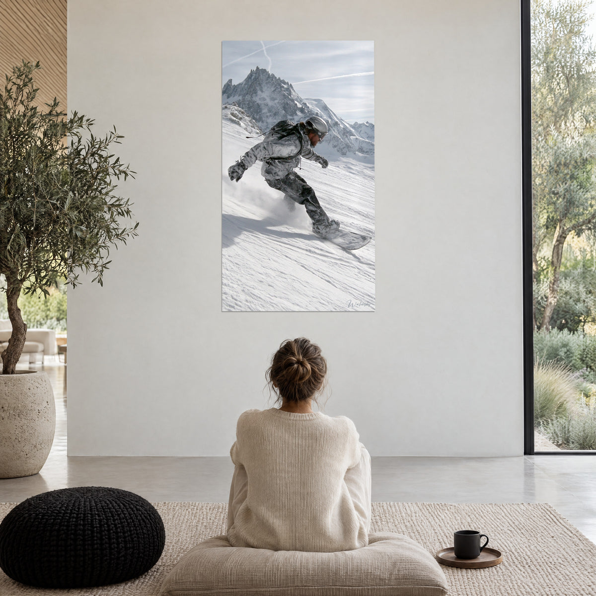Wall art snowboard in full descent, trail of white powder and steel-grey massif under a pale sky, facing a person seated with their back turned on a pouf