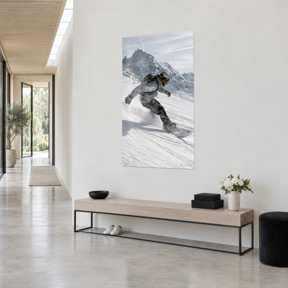 Wall art rider leaning into white powder snow, charcoal trousers and grey backpack, jagged peak behind, three-quarter view on a light wall