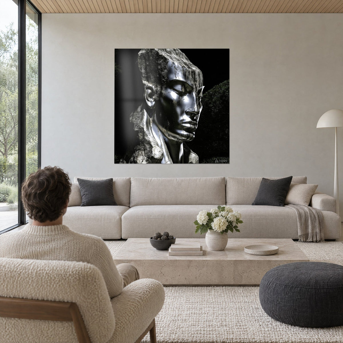 Quadro volto cromato scolpito nella roccia, riflessi neri e argento, uomo di spalle, sopra un divano in un soggiorno luminoso