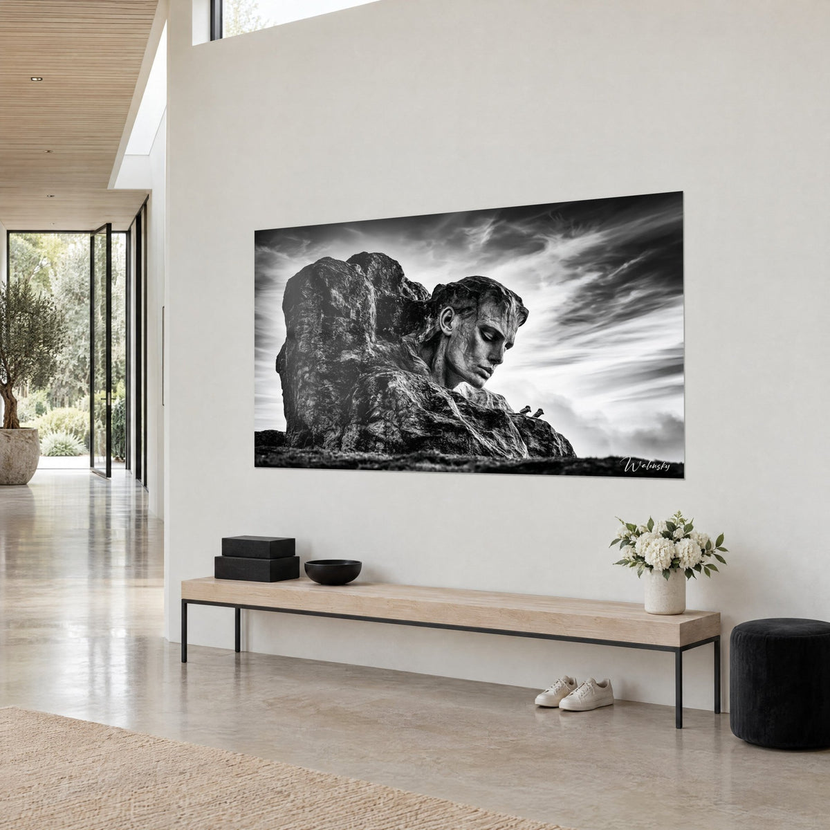 Tableau sculpture au profil penché, front de granit gris et nuages blancs filant, accroché sur un mur beige près d'une banquette en bois